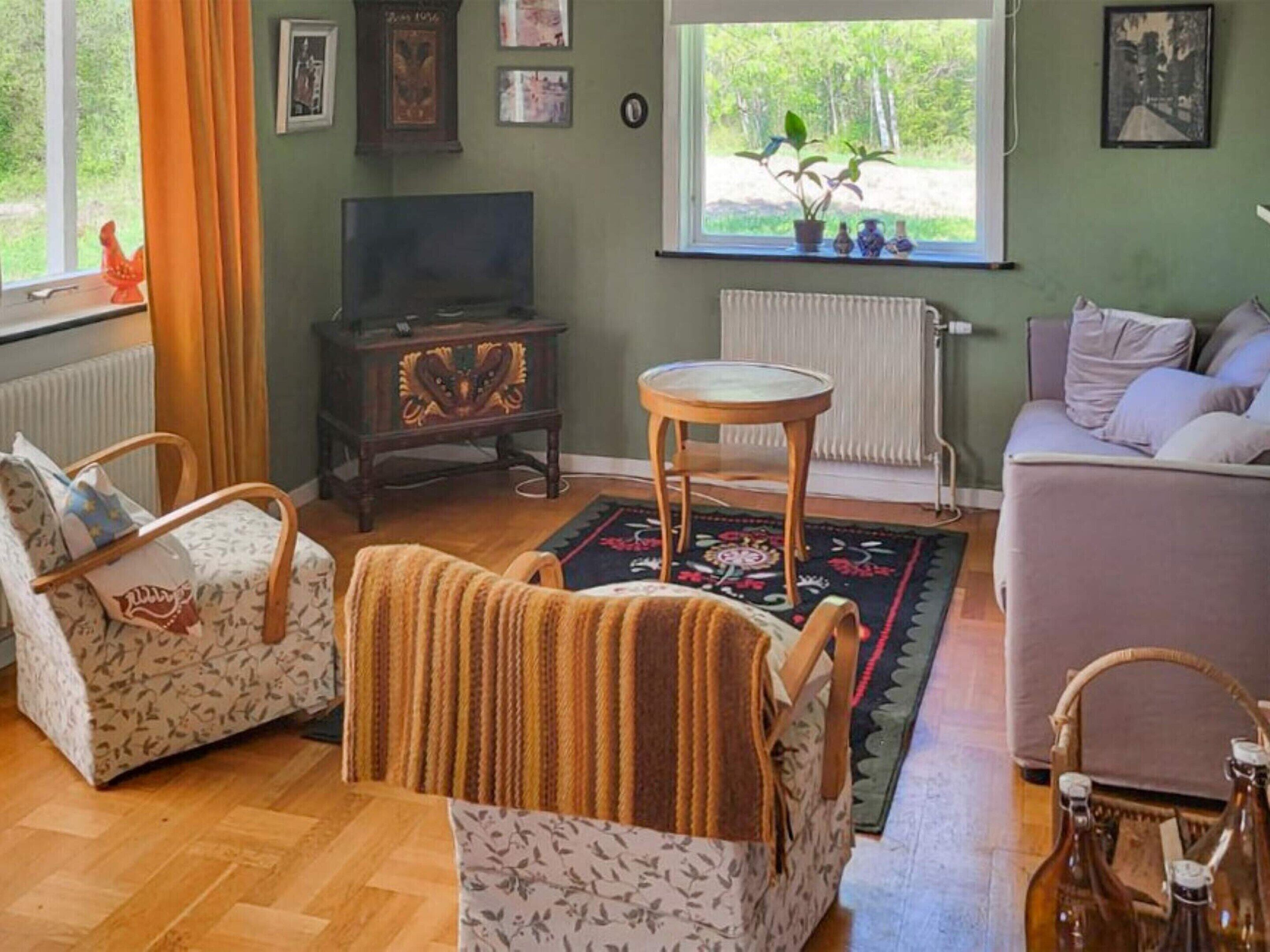 A cozy living room with a sofa, armchairs, a television, a small table, and a window with a view of greenery.