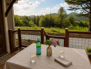 Un tavolo con una bottiglia, un vaso di fiori e dei bicchieri su un balcone affacciato su un paesaggio boschivo.