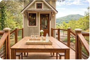 Un molo di legno con un tavolo apparecchiato per due, una piccola casa con balcone e un paesaggio montuoso sullo sfondo.