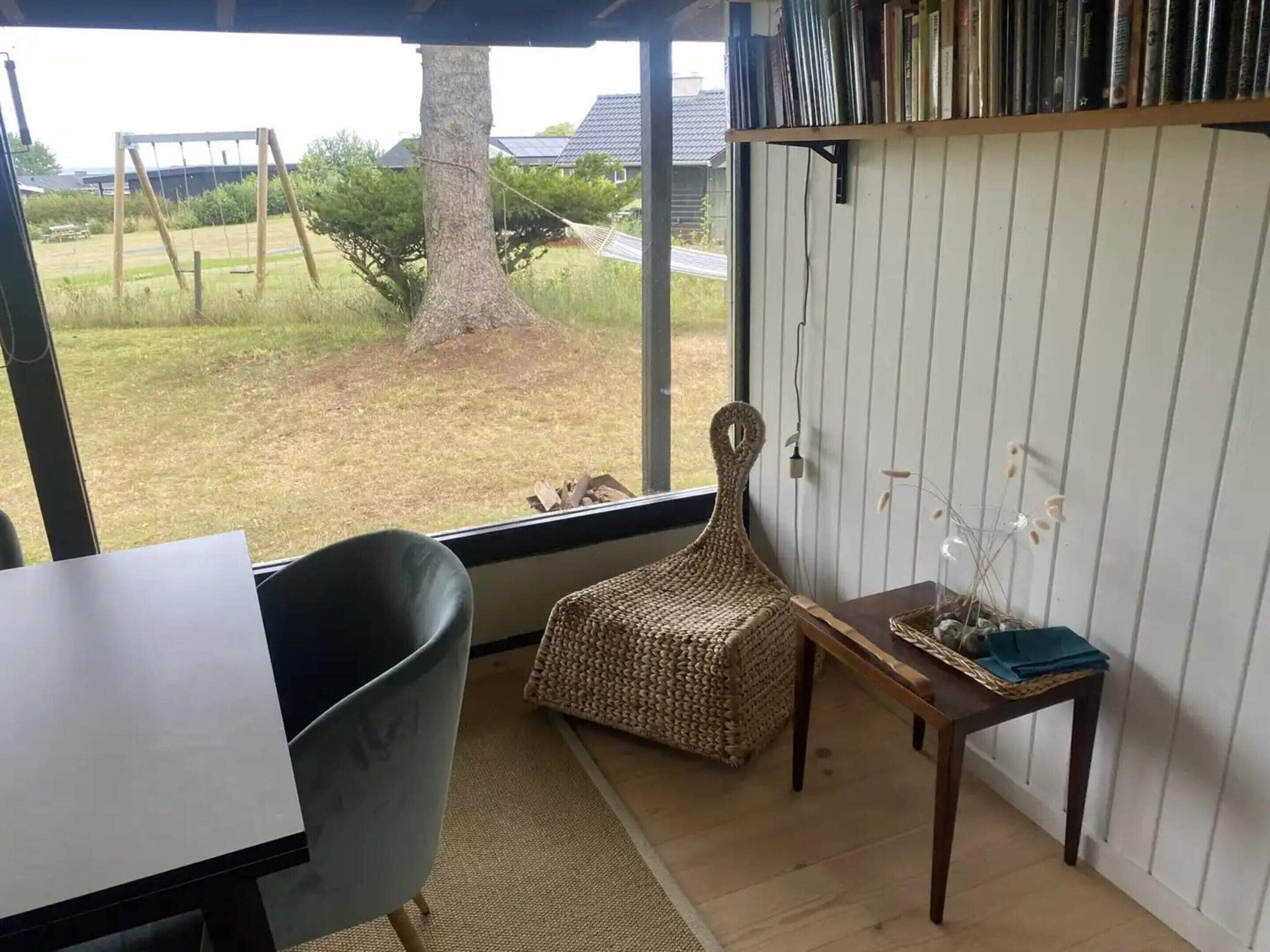 Una habitación con vistas a un espacio verde, un conjunto de columpios, una silla de mimbre, una mesita con un florero y una estantería.