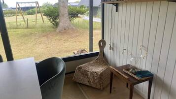 Una habitación con vistas a un espacio verde, un conjunto de columpios, una silla de mimbre, una mesita con un florero y una estantería.