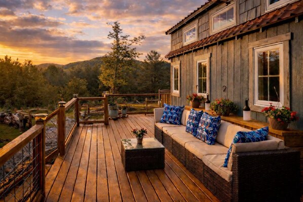 A wooden deck with a wicker sofa and potted plants, overlooking a forested area.