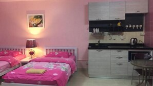 Un dormitorio con dos camas, una mesita de noche, una lámpara, un cuadro, una kitchenette con armarios y una pequeña mesa de comedor.