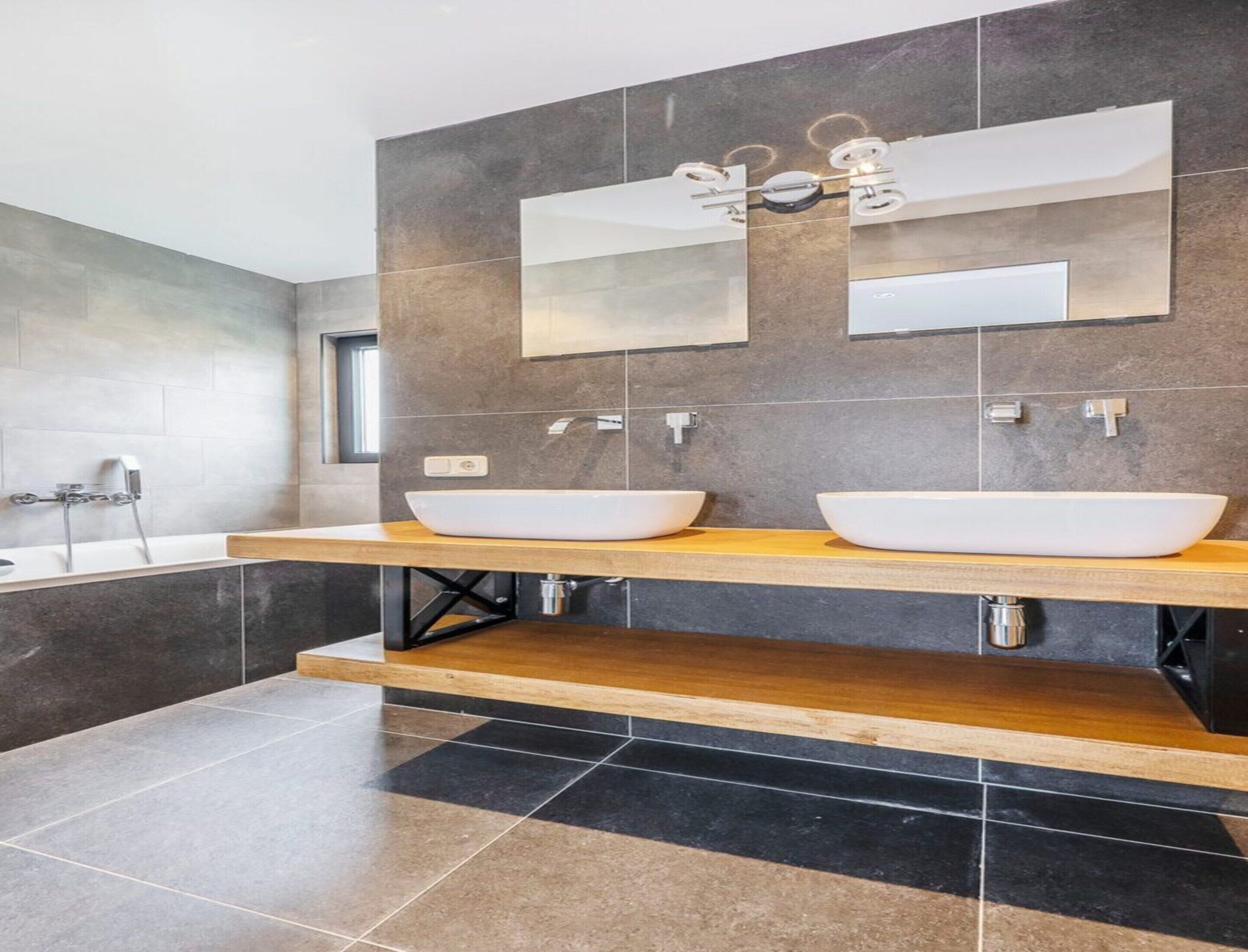 A modern bathroom with two large white sinks, a wooden vanity, a bathtub, and a large mirror above the sinks.