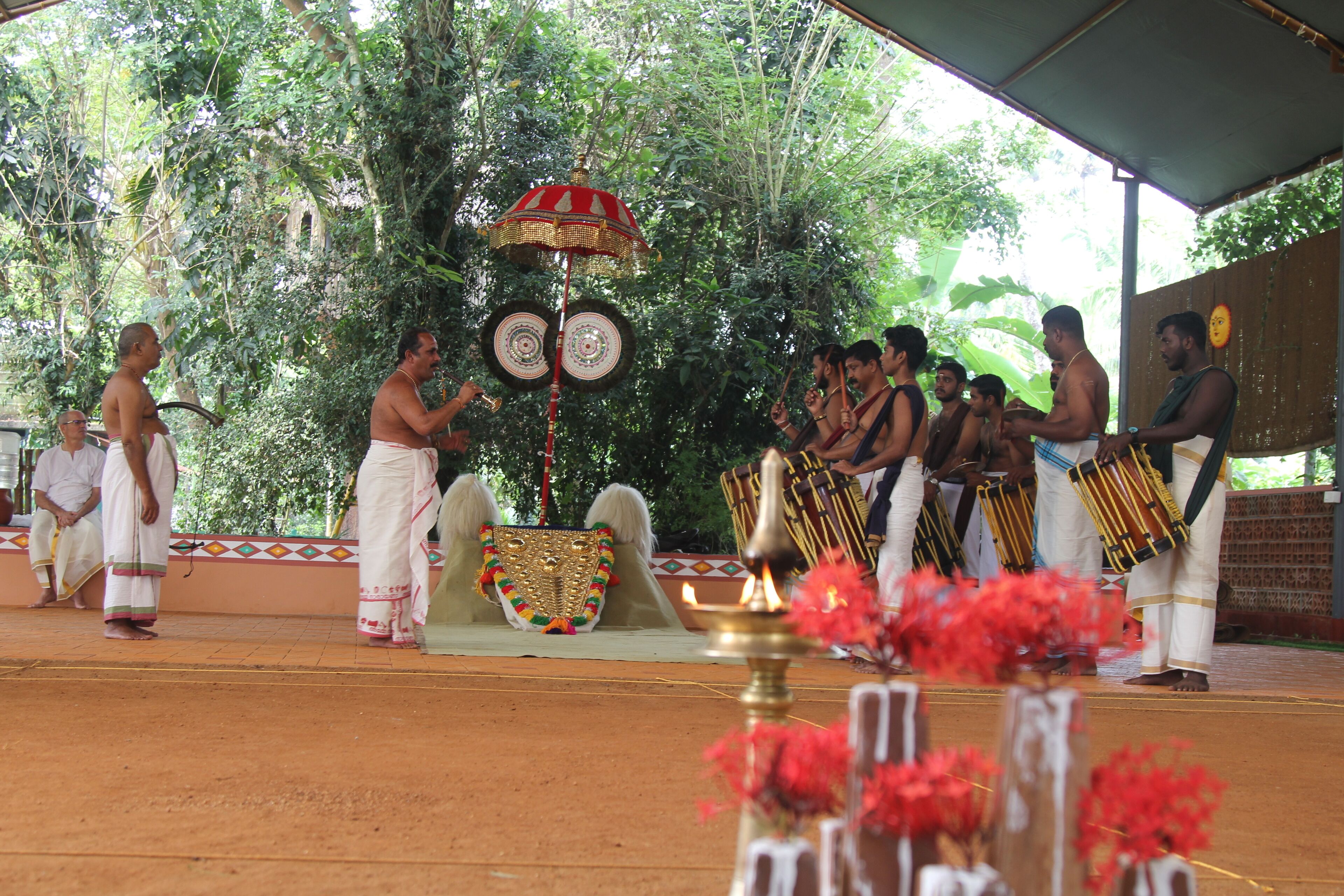 A group of people in traditional attire performing on a stage with musical instruments, surrounded by greenery.