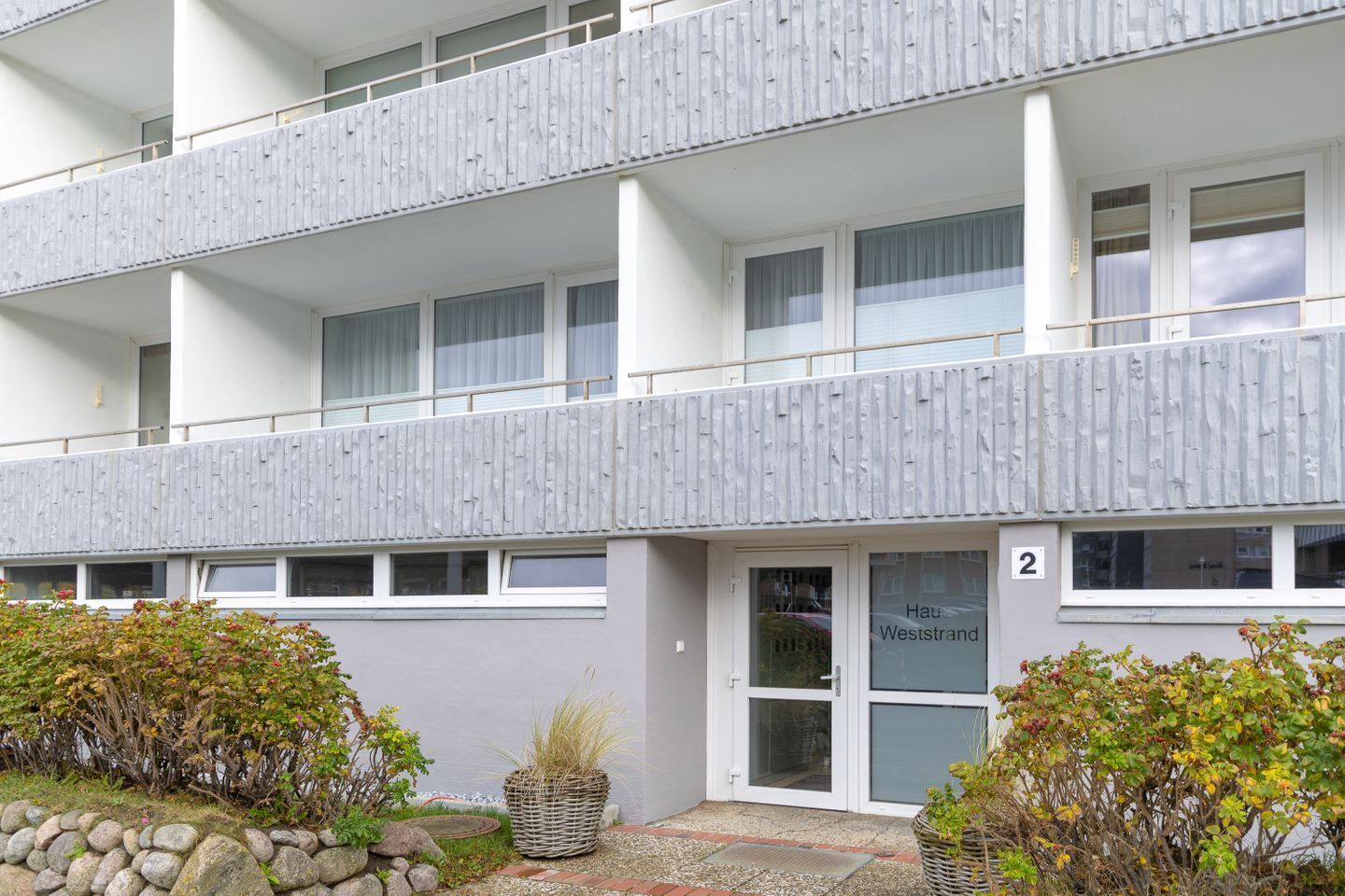 Un edificio de varios pisos con fachada plana, un balcón en el piso superior y una entrada a nivel de calle con una puerta marcada con el número '2'.