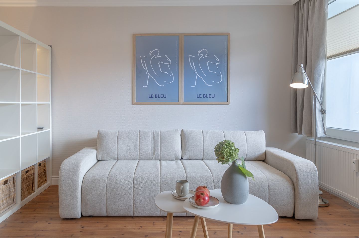 A modern living room with a grey sofa, a small round table with a vase and a pomegranate, and two framed artworks on the wall.
