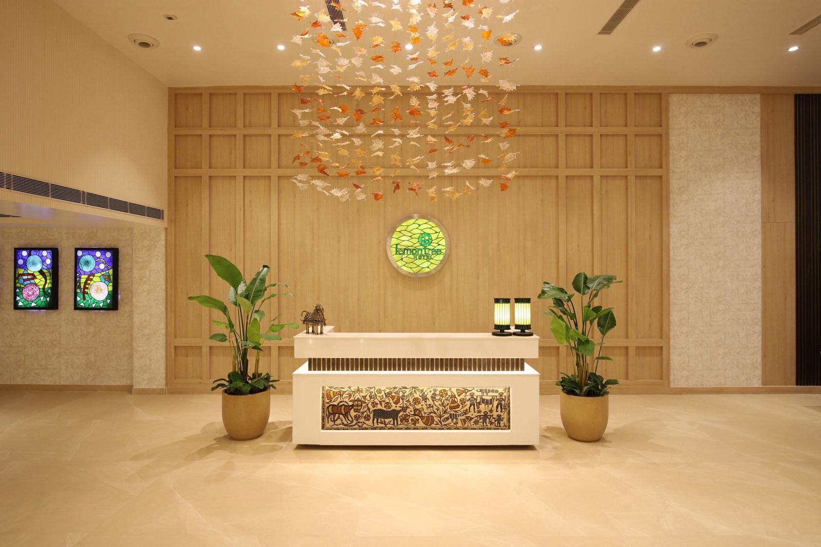 A reception area with a wooden panel wall, a central desk, two potted plants, a stained glass artwork, and two wall-mounted screens.