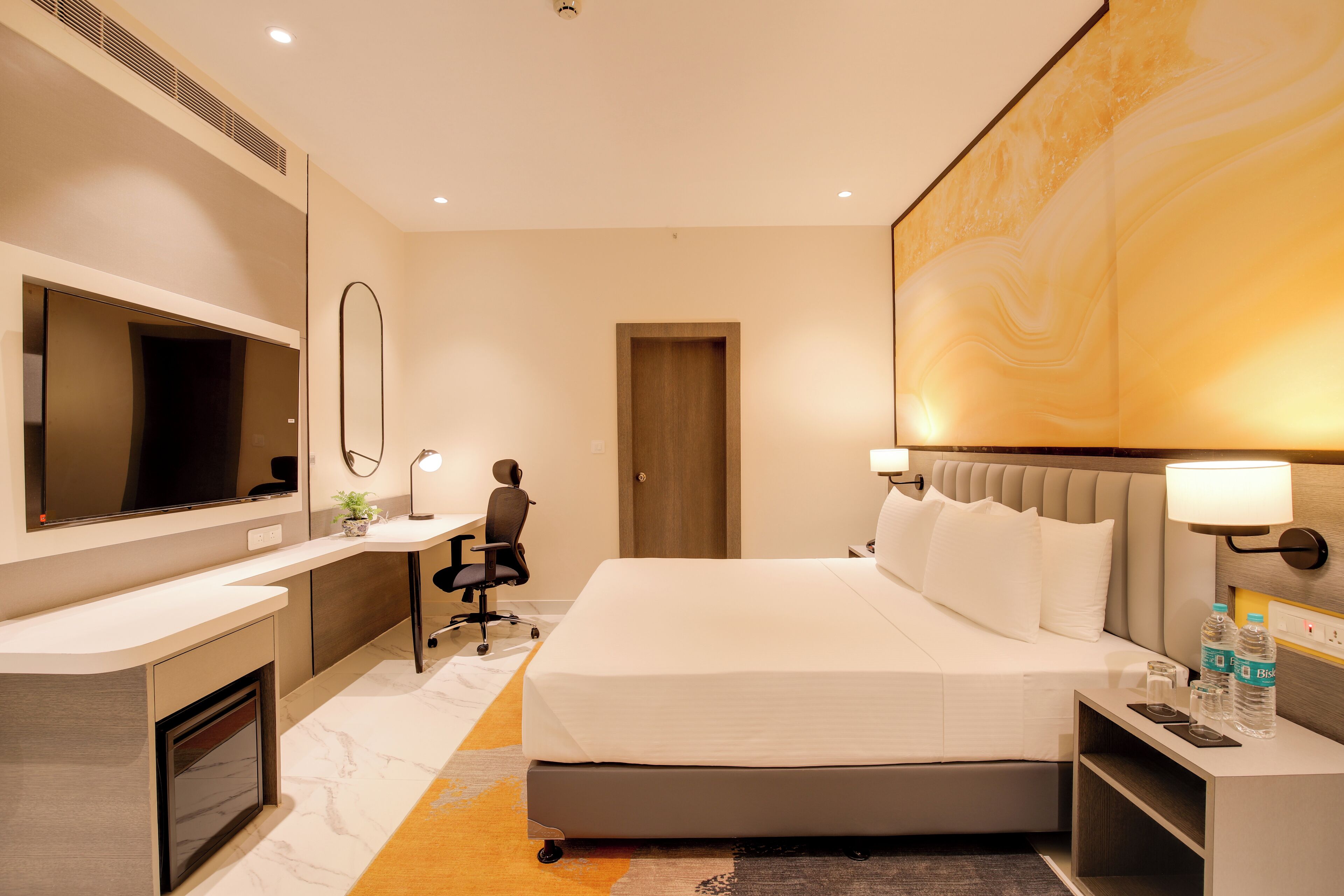 Una habitación de hotel con una cama, un escritorio, una silla, un televisor y un espejo grande.