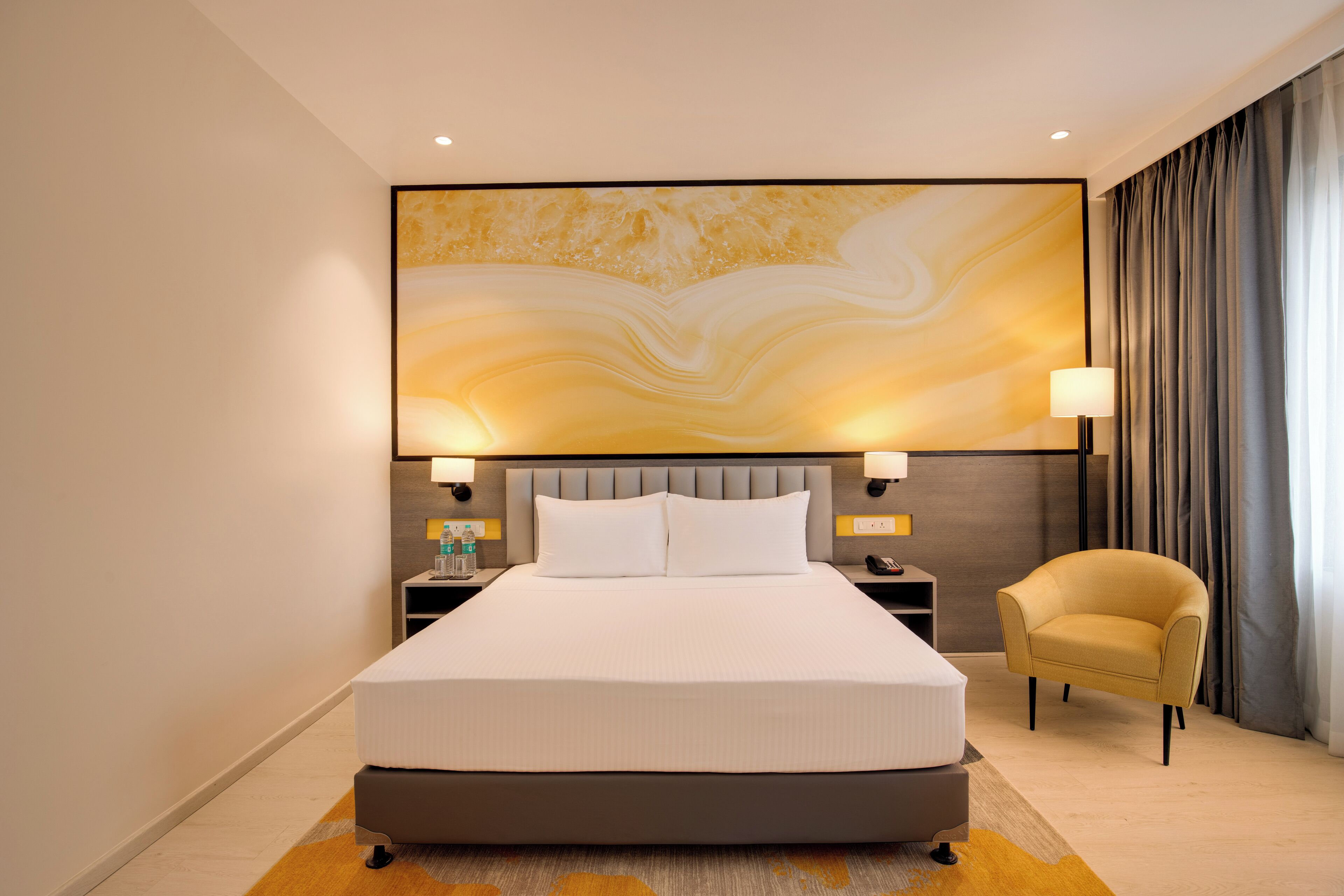 Une chambre d’hôtel moderne avec un grand lit, une tête de lit au motif évoquant le marbre, deux lampes de chevet, une table de chevet, une chaise et un grand tableau abstrait accroché au mur.