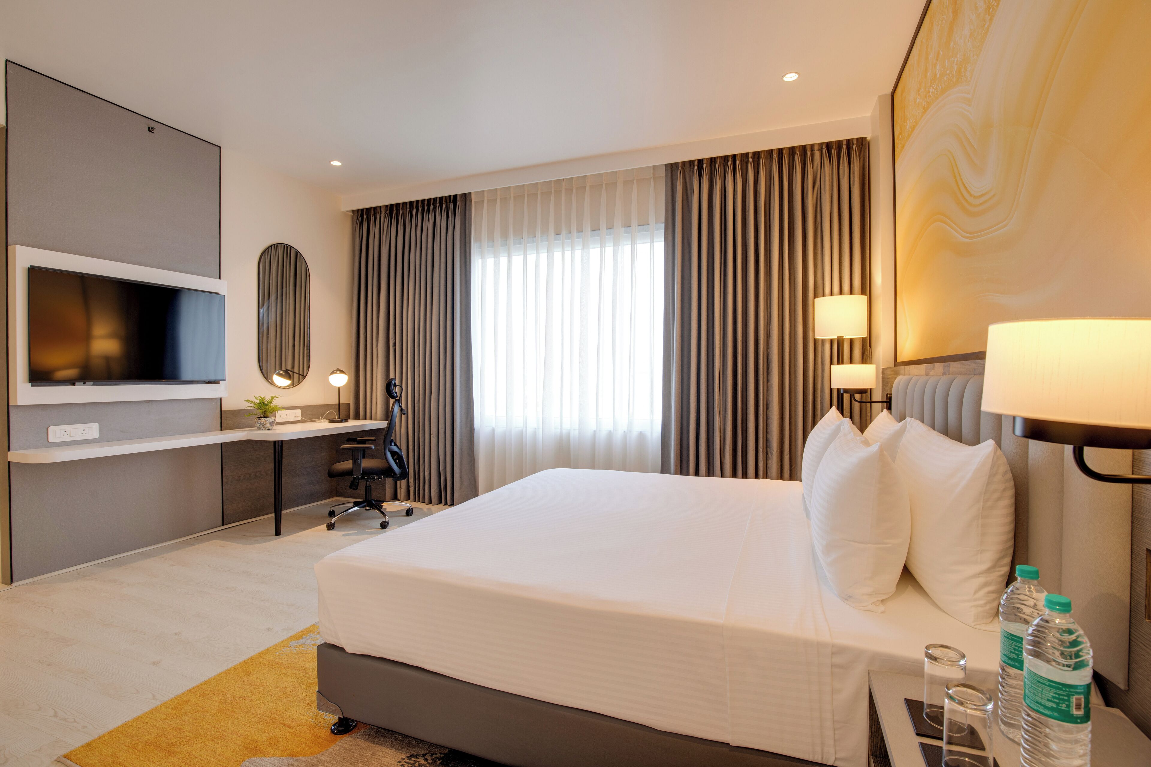 A modern hotel room with a large bed, a desk with a chair, a flat-screen TV, and a large window with curtains.