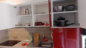 A compact kitchen with a sink, a stove, and a red cabinet with open shelves containing kitchenware.