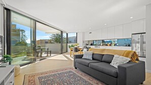 A modern apartment with a large sliding glass door leading to a balcony, a cozy living area with a sofa and a patterned rug, and a kitchen with white cabinets and a stainless steel refrigerator. - ROCKPOOL #101 | Walk to beach, WIFI | Accom Holidays (TERRIGAL)