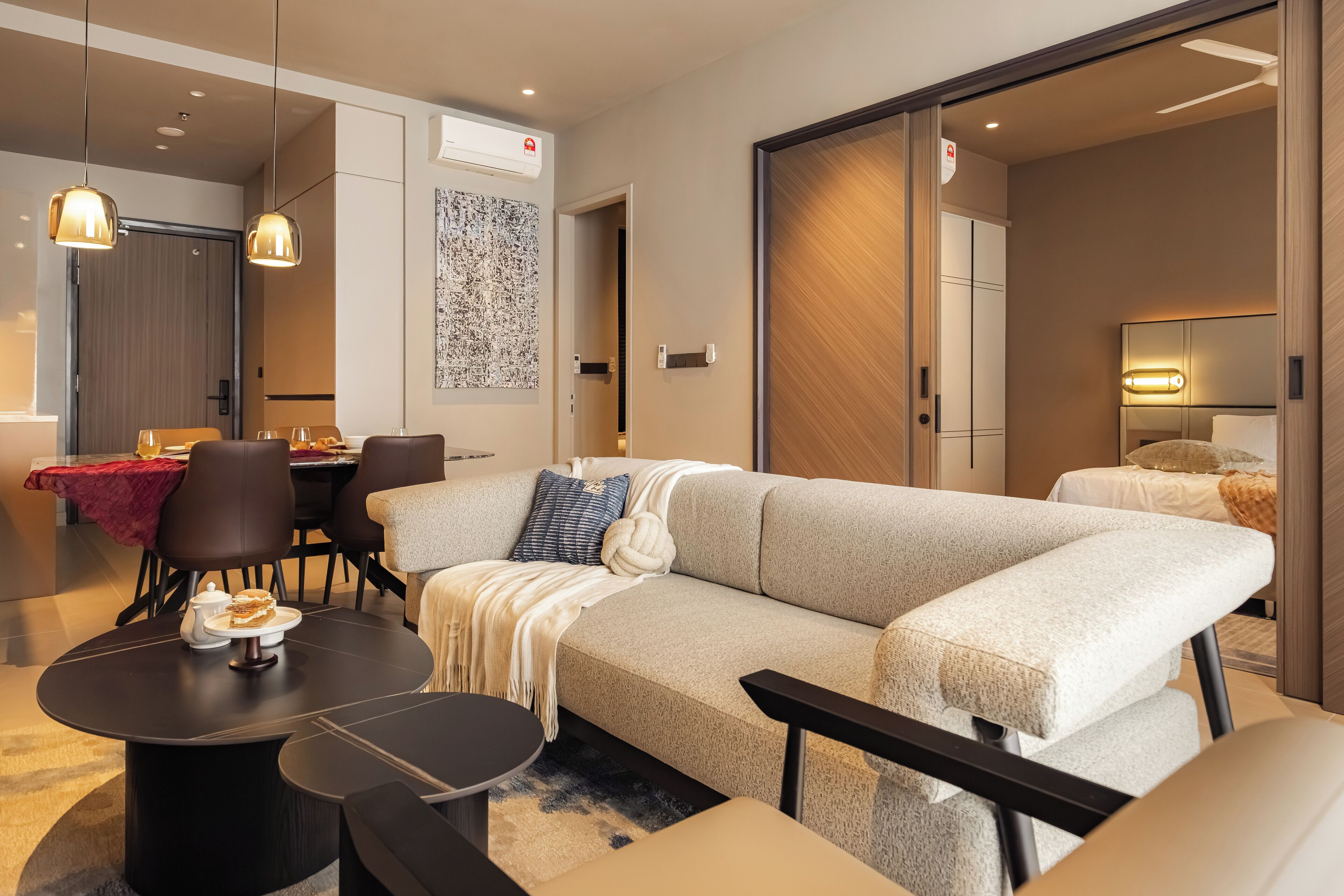 A modern hotel room with a large beige sectional sofa, a dining table with chairs, a round coffee table, and a separate seating area with a bench.