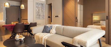 A modern hotel room with a large beige sectional sofa, a dining table with chairs, a round coffee table, and a separate seating area with a bench.