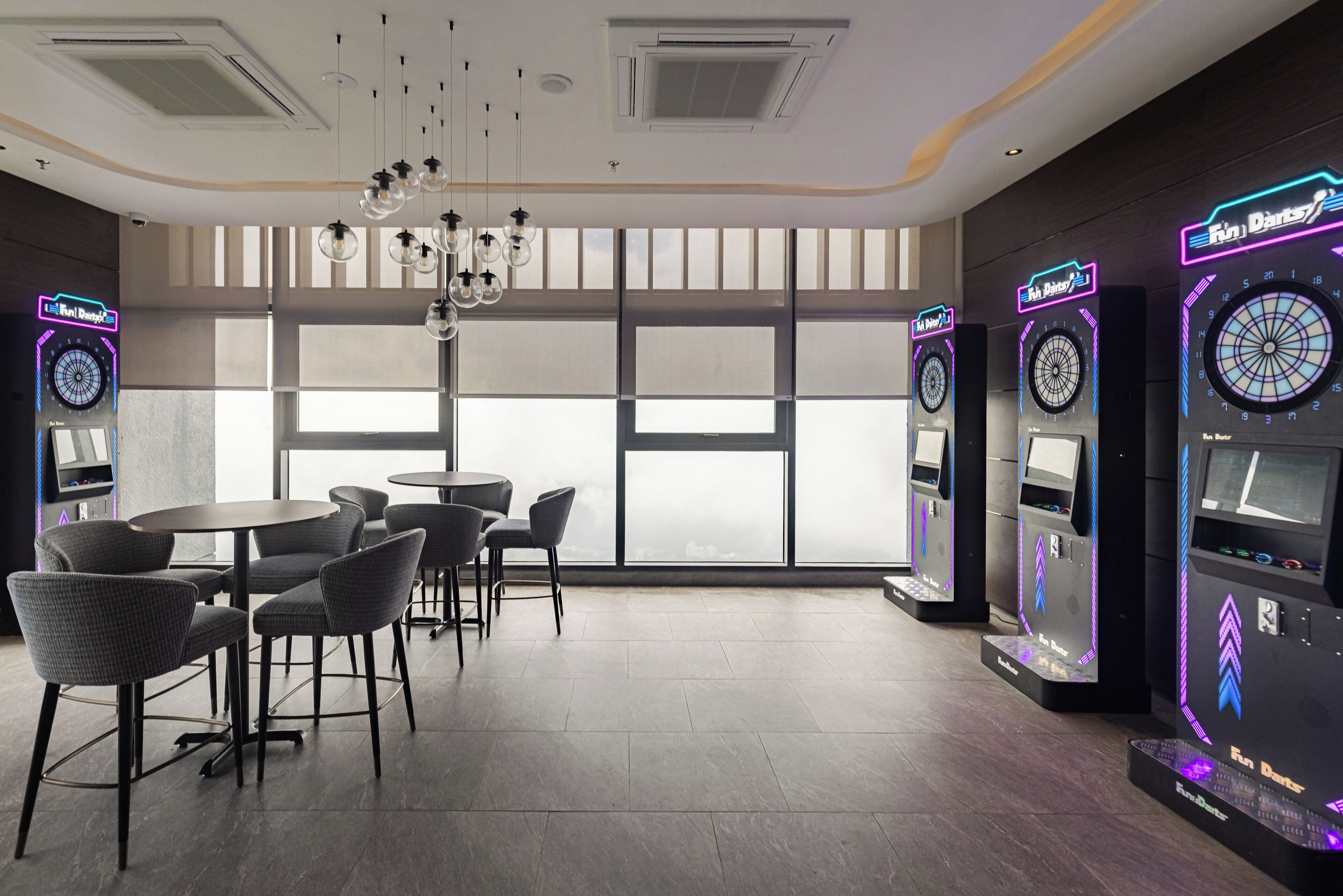 A modern indoor space with tables and chairs, large windows, and dartboards.