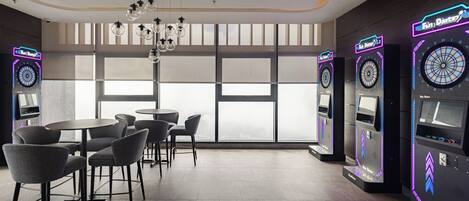 A modern indoor space with tables and chairs, large windows, and dartboards.