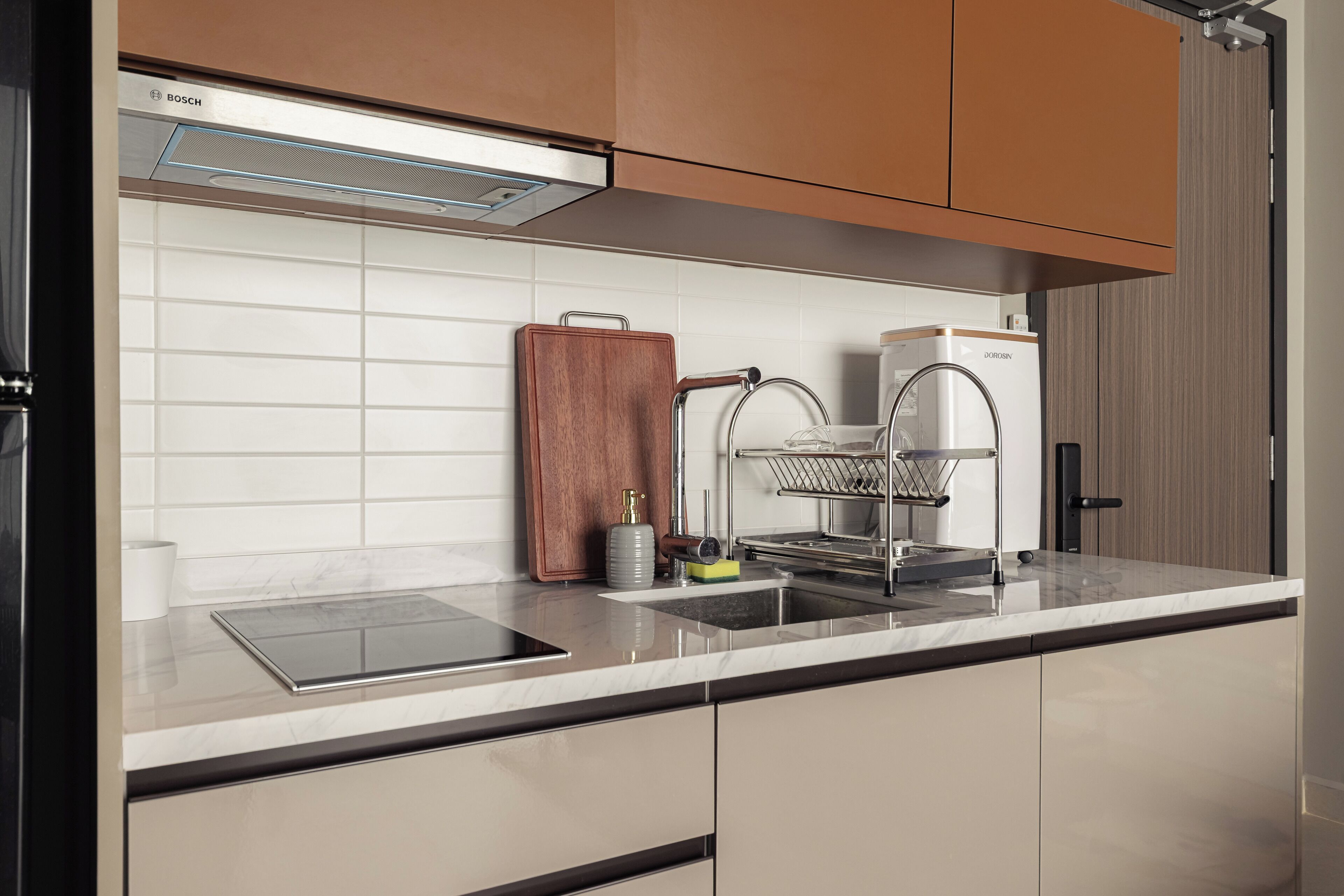A modern kitchen with a Bosch range hood, a wooden cutting board, a sink with a dish rack, and a refrigerator.