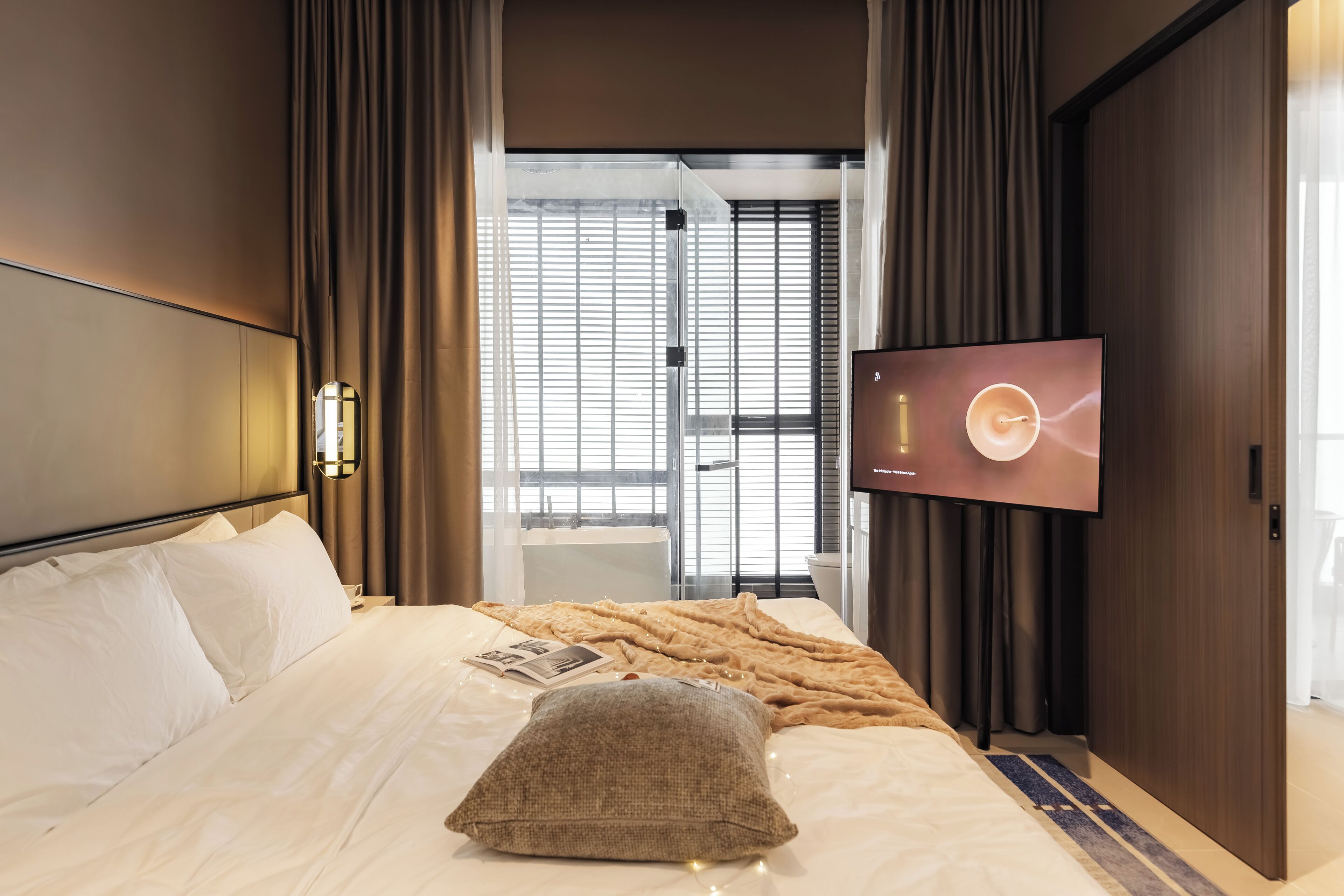 A hotel room with a bed, a television, and a view of the outside through a window with blinds.