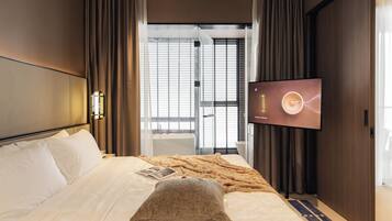A hotel room with a bed, a television, and a view of the outside through a window with blinds.