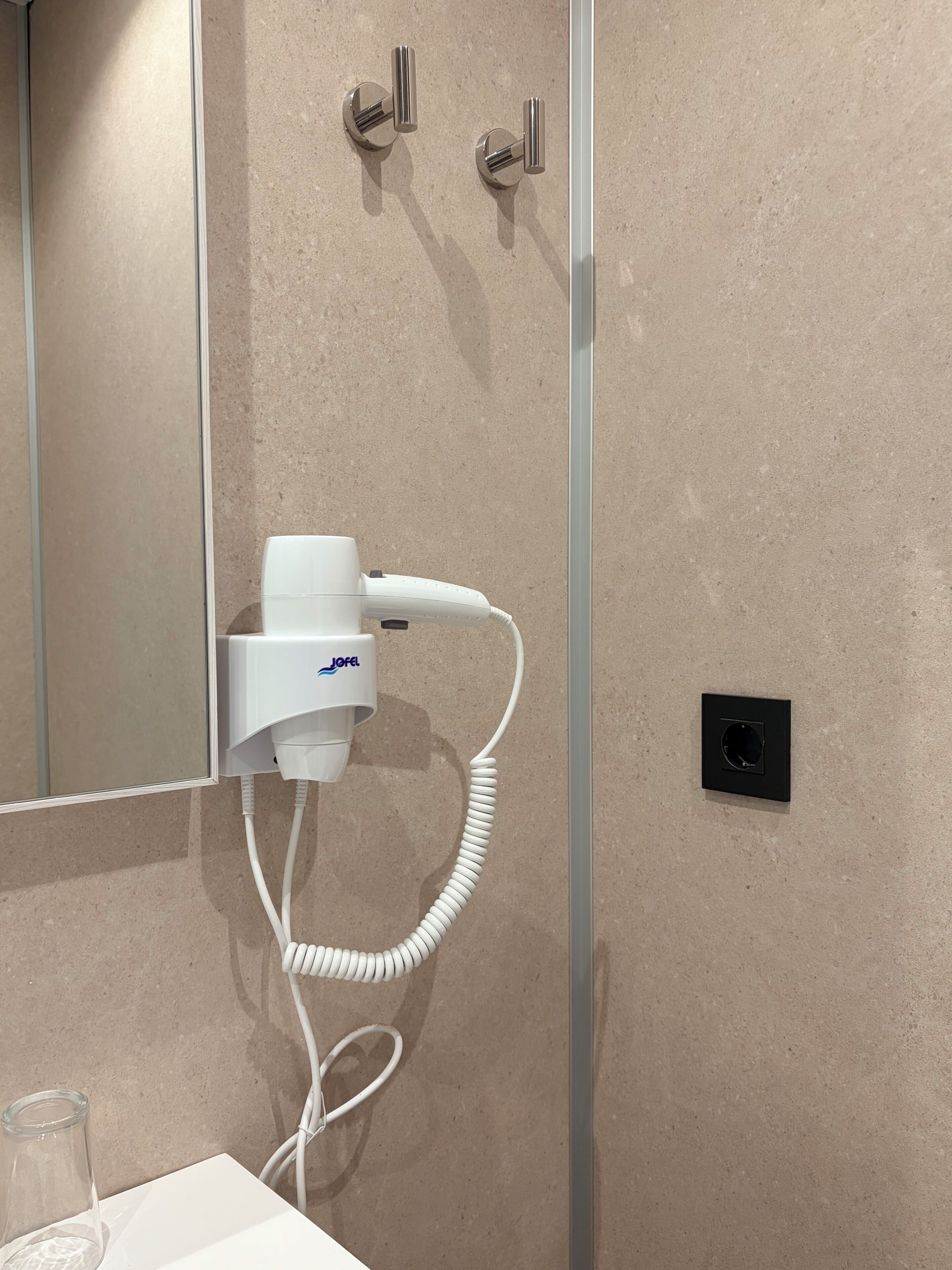 Un baño con un secador de pelo montado en la pared, un espejo y un vaso de agua sobre el mostrador.