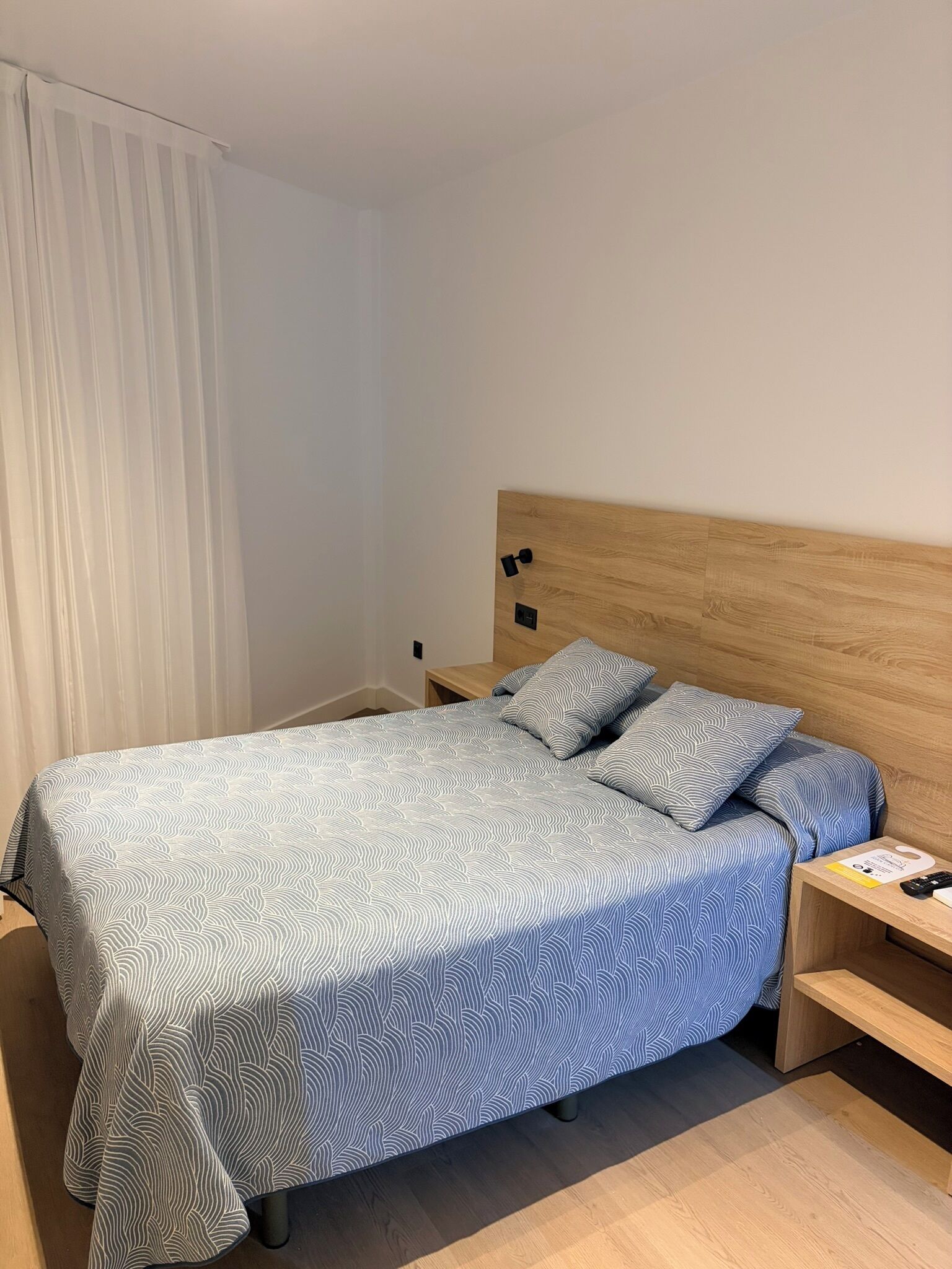 Una habitación de hotel con cama, mesita de noche, cabecera de madera y ventana con cortinas blancas.