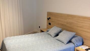 A hotel room with a bed, a nightstand, a wooden headboard, and a window with white curtains.