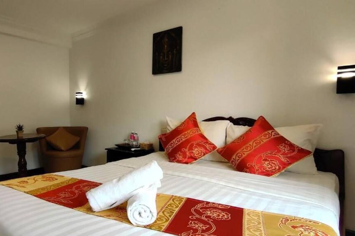 Una habitación de hotel con una cama bien hecha, dos sillones, una mesita redonda y una lámpara fijada a la pared.