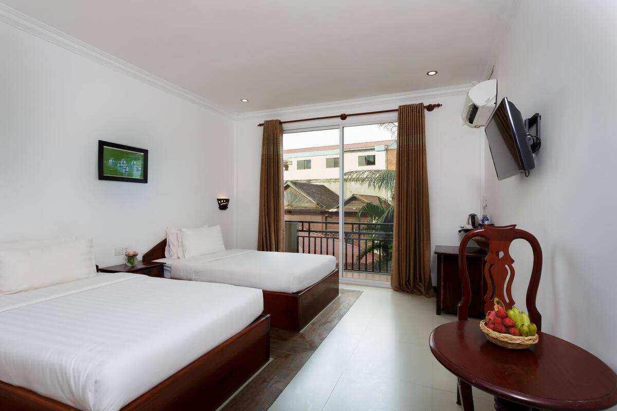Habitación de hotel con dos camas, un televisor montado en la pared, un balcón con cortinas y una mesita con una cesta de fruta.