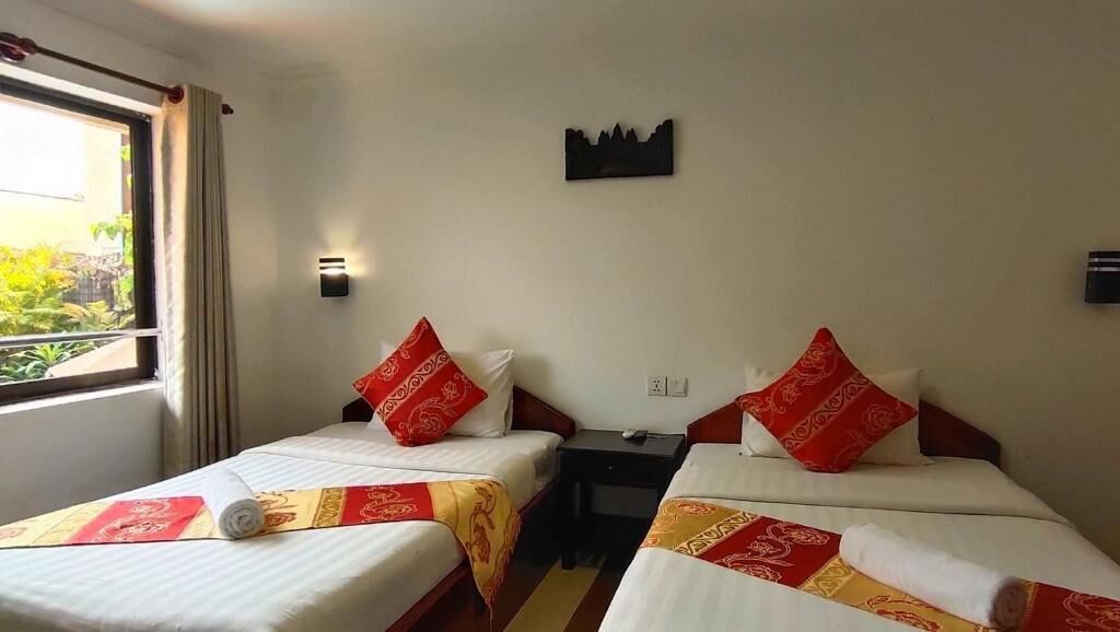 Ein Hotelzimmer mit zwei Betten, jedes mit einem roten Kissen und einer weißen Decke. Dazwischen steht ein Nachttisch, und auf der linken Seite befindet sich ein Fenster mit Vorhängen.