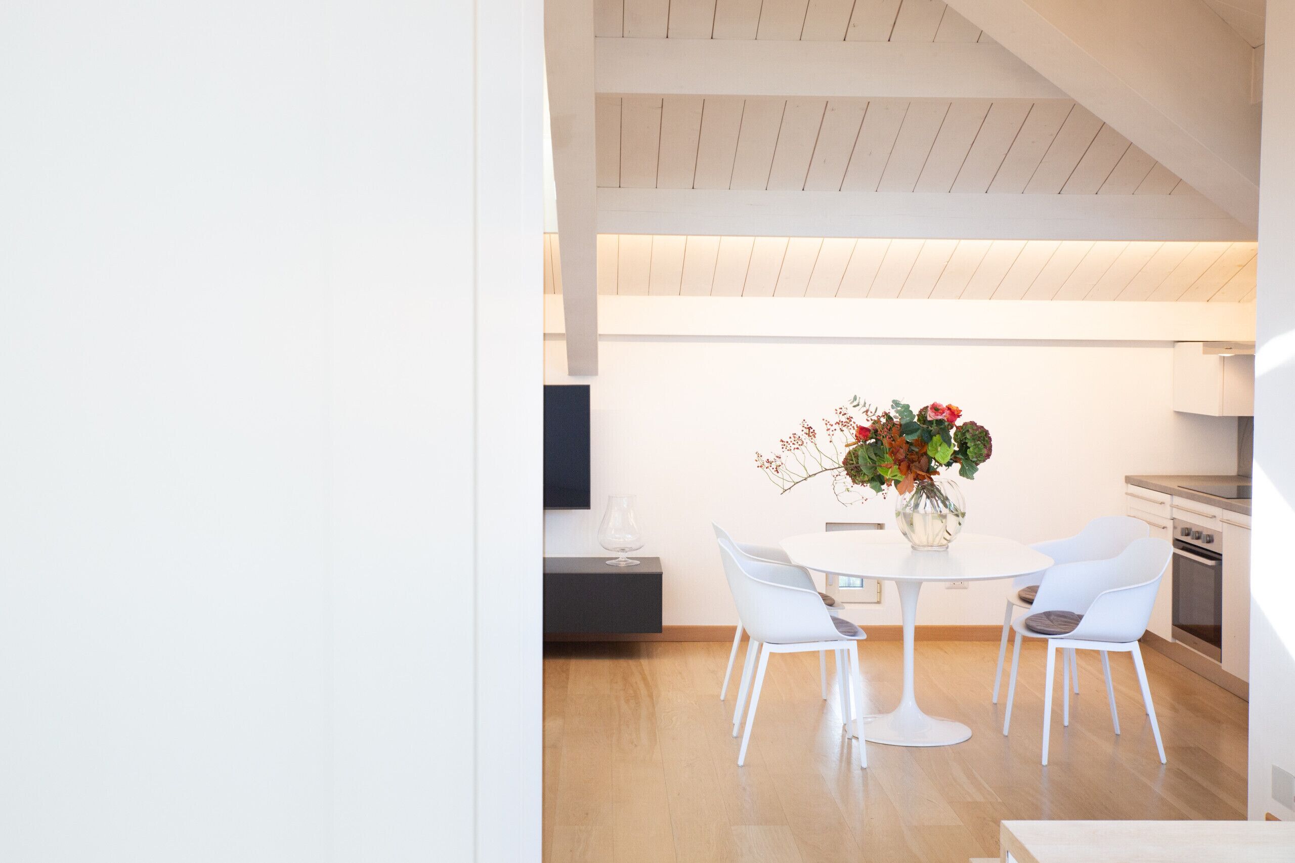 A modern kitchen with white cabinetry, a dining area with a round table and white chairs, and a wooden ceiling.