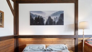 Een kamer met een houten hoofdbord, twee bedden met lichtgekleurde beddengoed, een lijst met daarop een afbeelding van een bos, en een bedlamp.