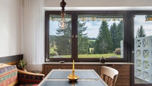 Een kamer met een groot raam, een tafel met een kaars, een stoel en een bank met kussens.