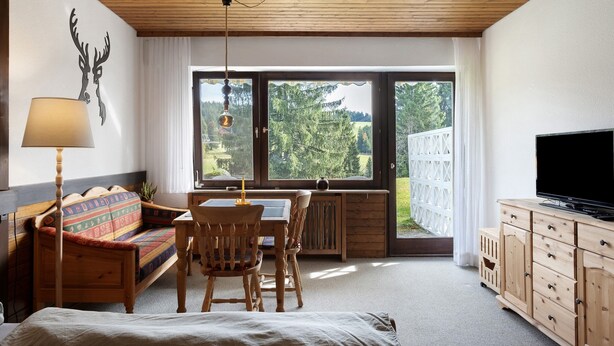 Een kamer met een bed, een houten bank, een eettafel, een televisie en een groot raam met uitzicht op bomen.