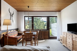 Una habitación con una cama, un sofá de madera, una mesa de comedor, un televisor y un ventanal con vistas a árboles.