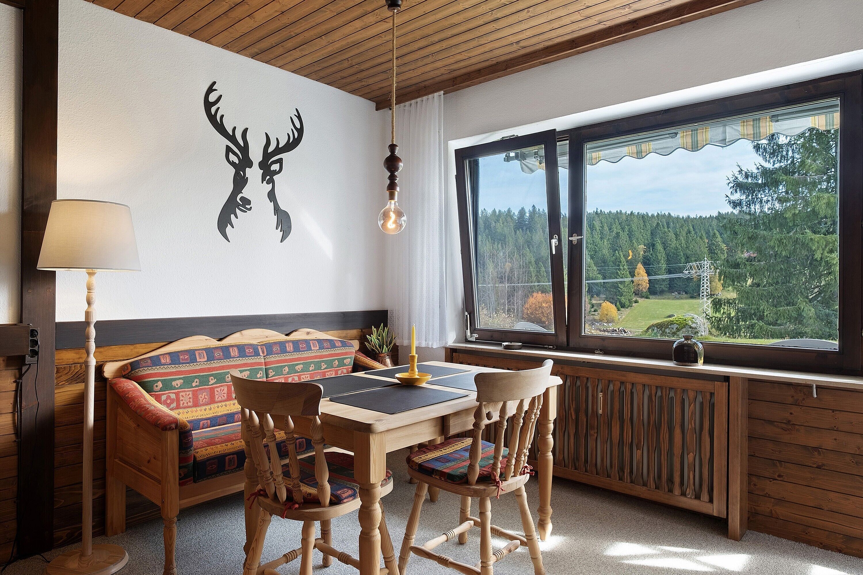 A cozy room with a bed, wooden furniture, a table, chairs, a lamp, a radiator, and a window with a view of trees.