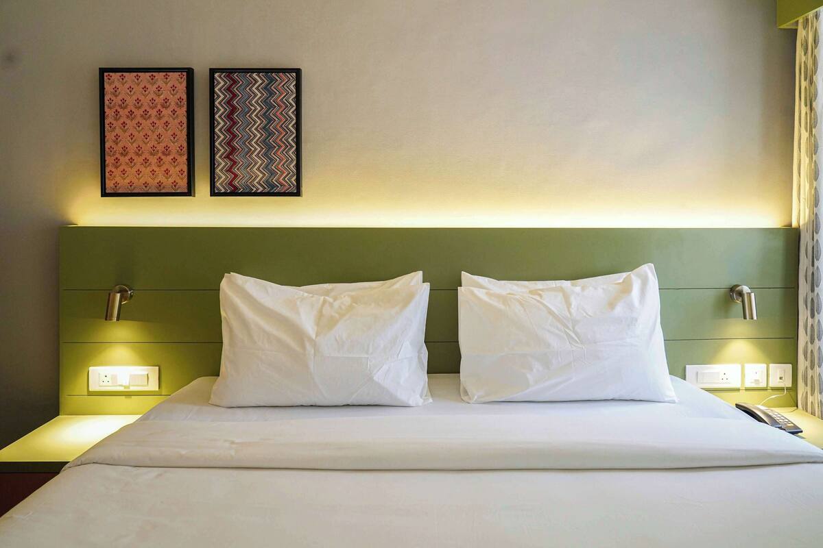 A hotel room with a bed, two white pillows, a green headboard, two wall-mounted nightstands, and two framed pictures on the wall.