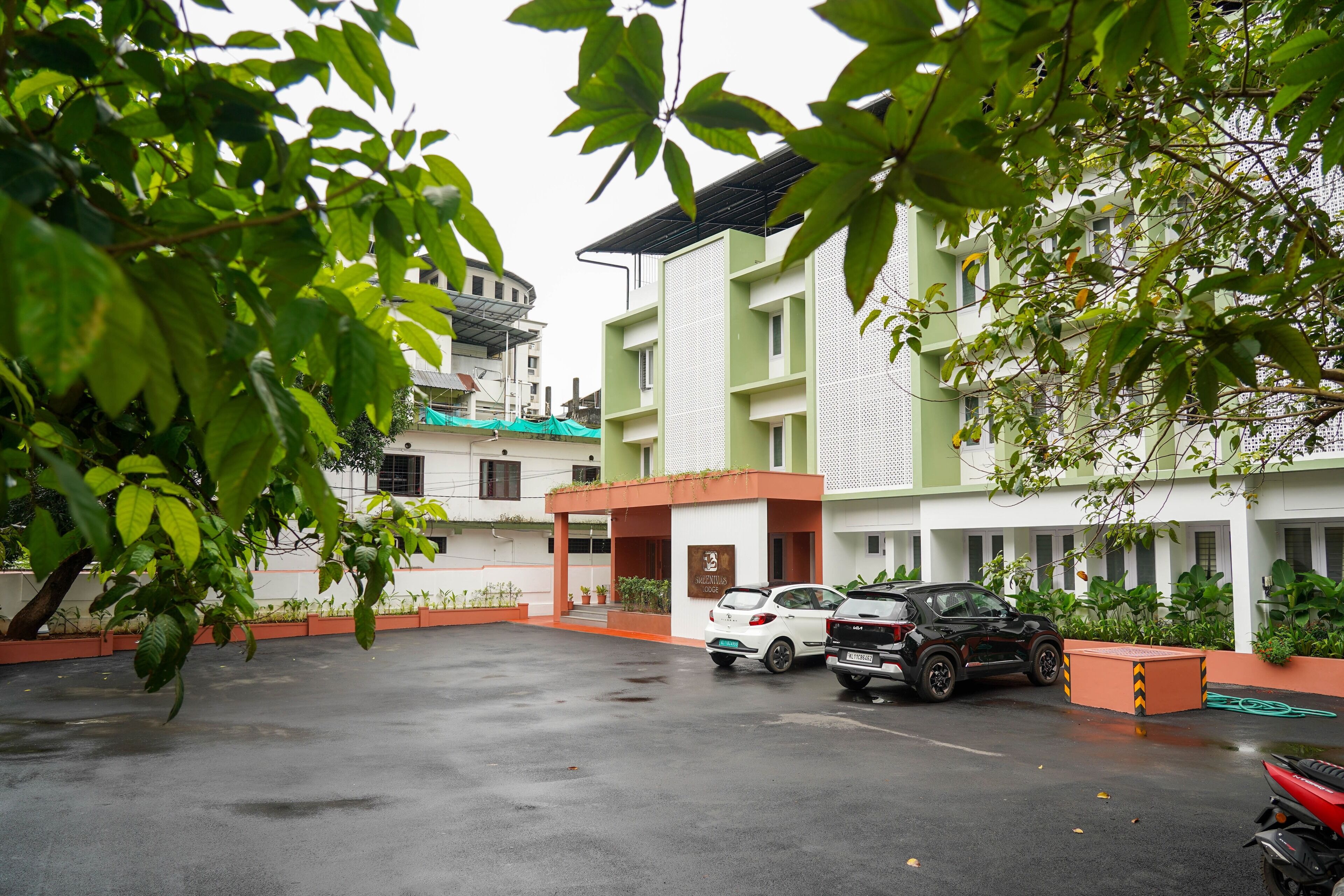A parking area with two cars and a motorcycle, surrounded by greenery and buildings with a modern design.