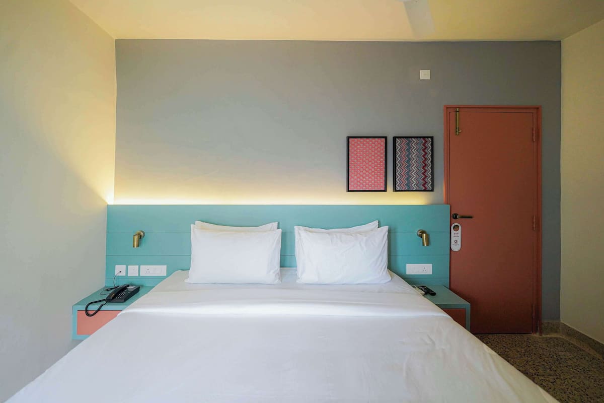 A hotel room with a bed, a nightstand with a phone, a door with a lock, and two wall-mounted pictures.