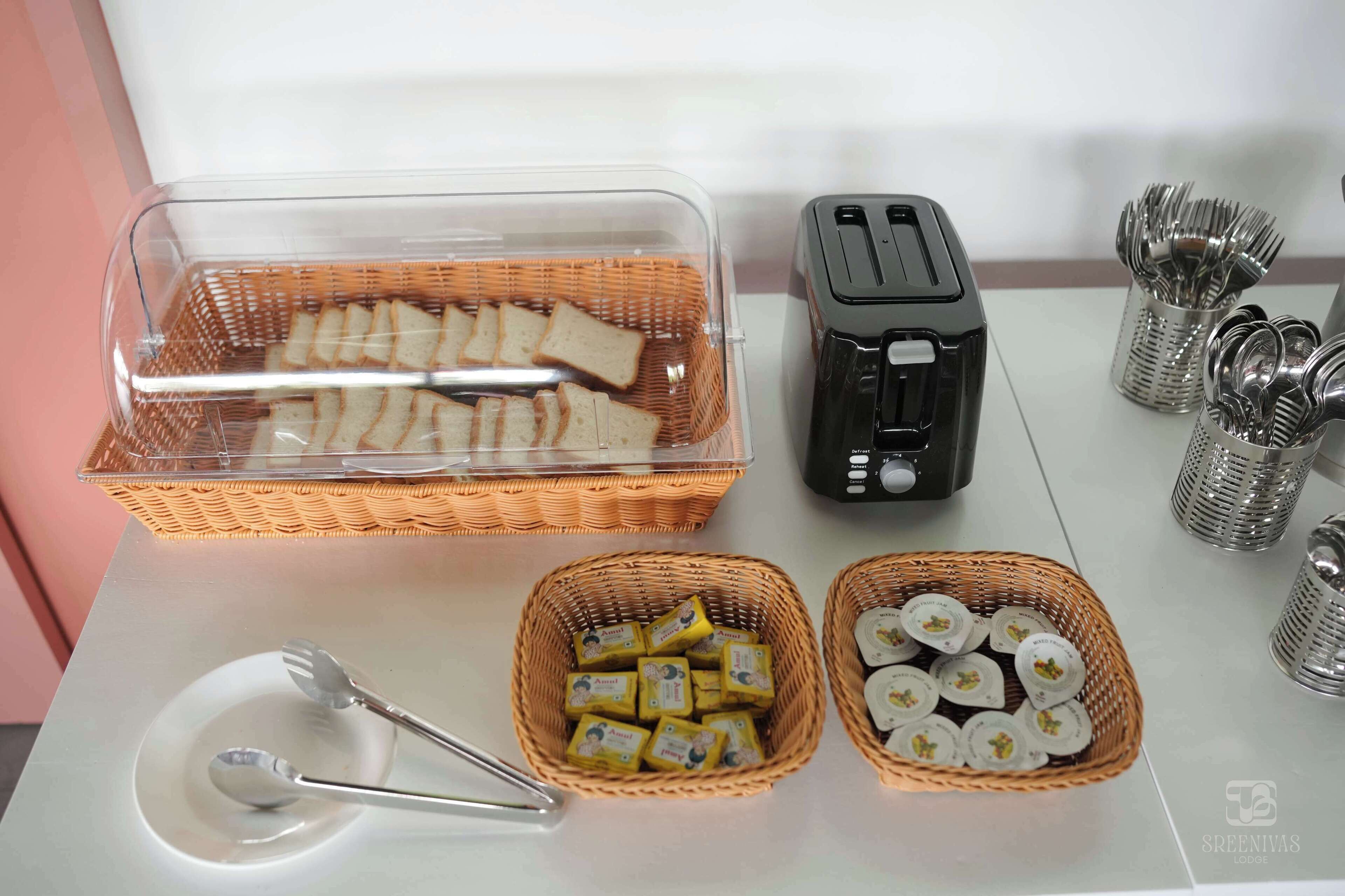 Disposição de buffet com recipiente plástico transparente contendo pão fatiado, uma torradeira e cestas com manteiga em sachês.
