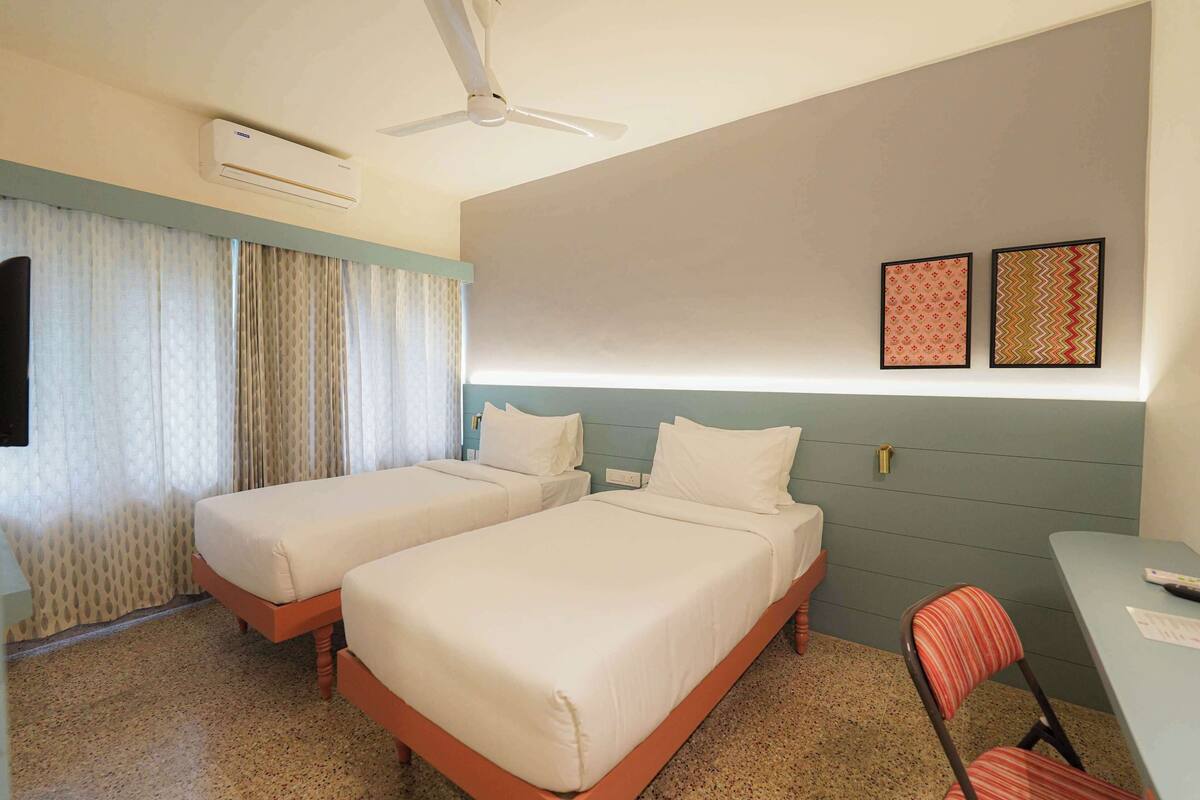 A hotel room with two beds, a desk, a chair, an air conditioner, a ceiling fan, and two framed pictures on the wall.