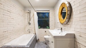 Un baño con bañera, inodoro y lavabo, con una pared de ladrillo y ventana con vistas a árboles.