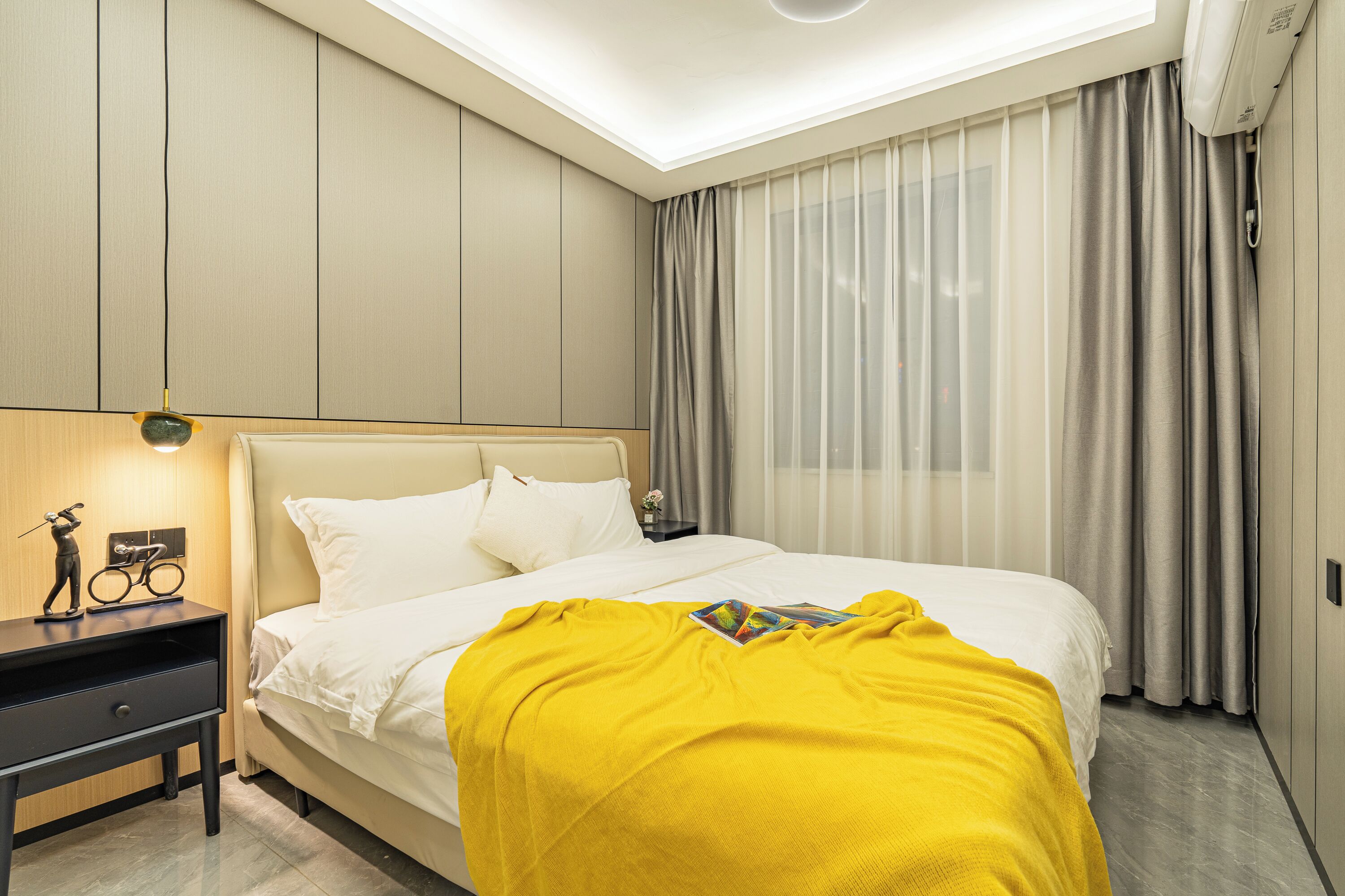 A modern hotel room with a large bed, a bedside table with a lamp, and a view of the city through sheer curtains.