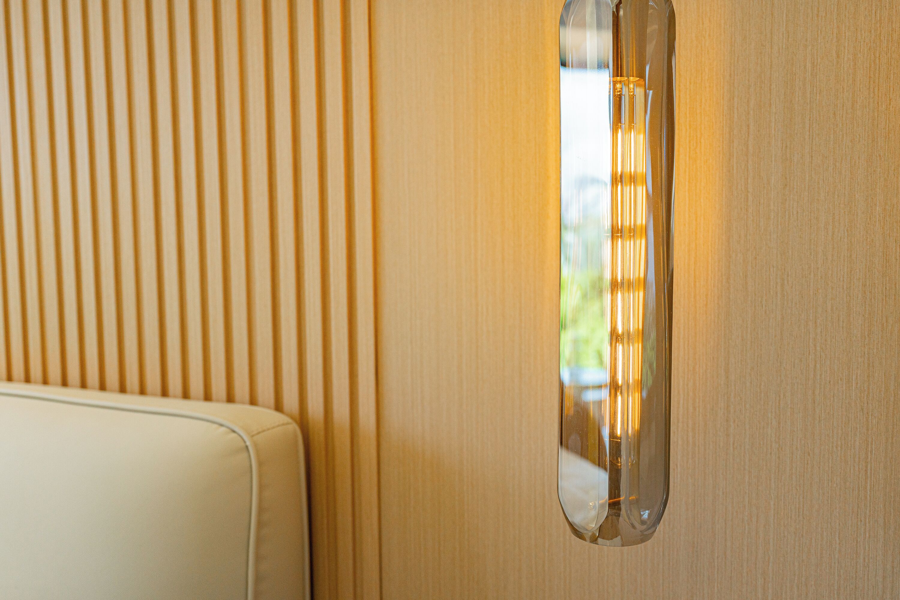 Una lámpara de pared con pantalla de vidrio transparente, un panel de pared de madera y un banco blanco acolchado.