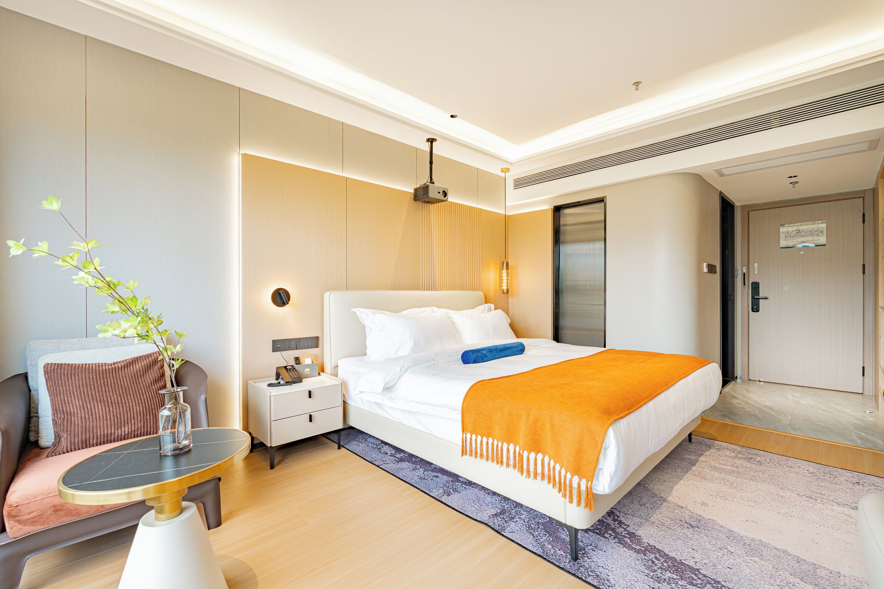 A modern hotel room with a neatly made bed, a bedside table with a lamp, a small table with a vase, and a chair.