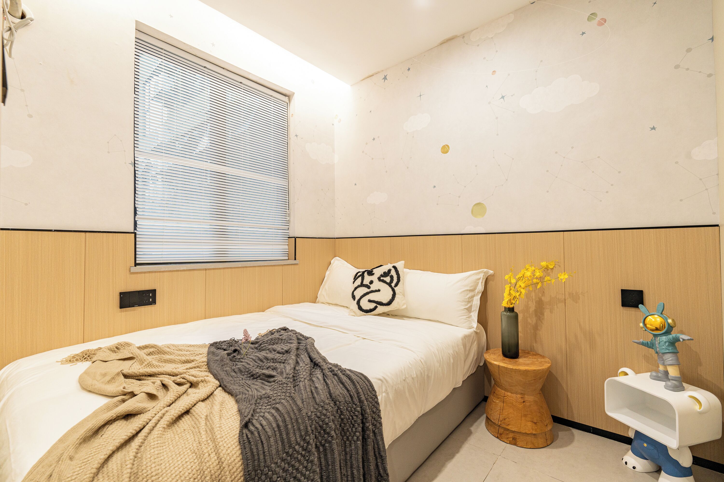 A bedroom with a bed, a window with blinds, a wall with a starry design, a wooden nightstand, and a small white nightstand with a toy figure.