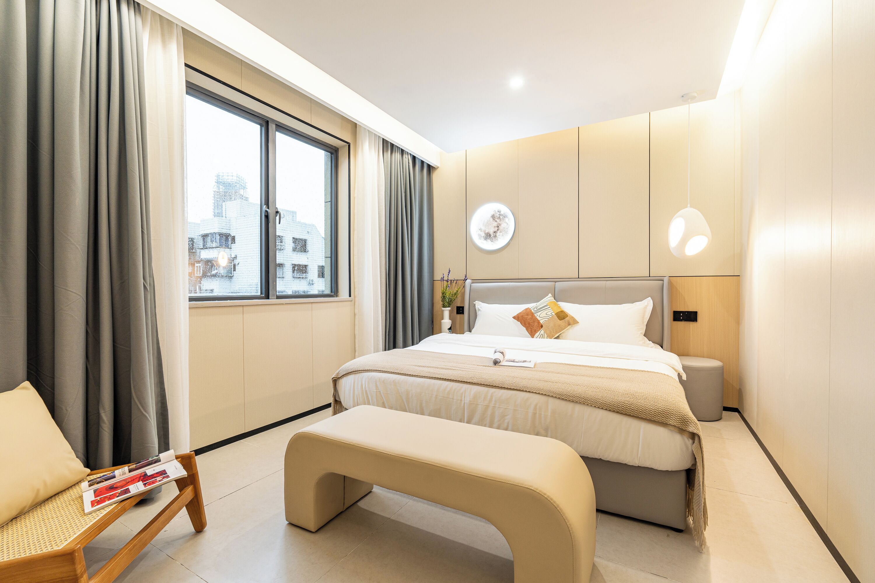 Ein modernes Hotelzimmer mit einem großen Bett, einer Bank, einem Nachttisch und Blick auf die Stadt durch ein großes Fenster.