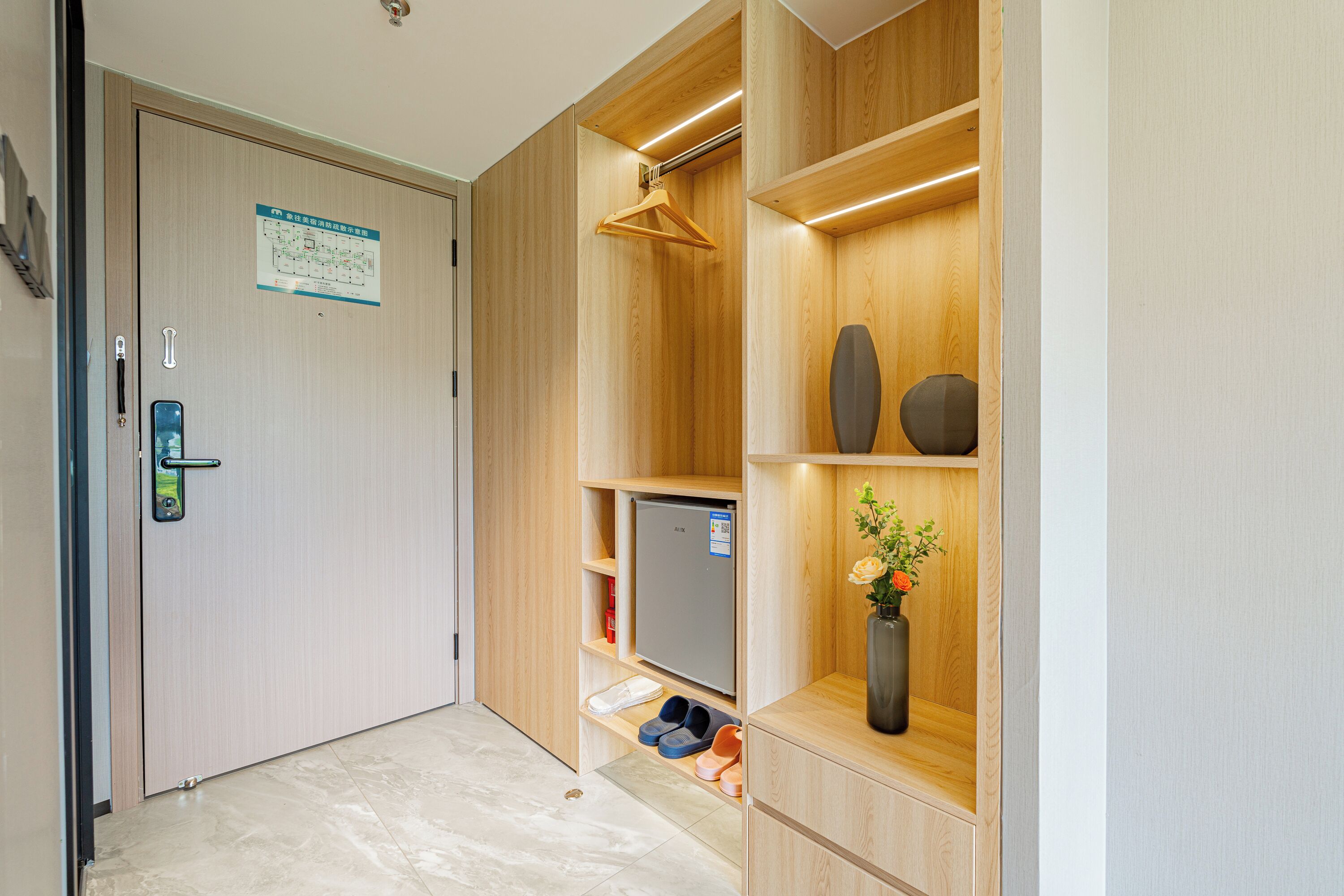 Une chambre d’hôtel avec une porte en bois, un porte-manteau, un petit réfrigérateur, un vase avec des fleurs et un porte-chaussures.