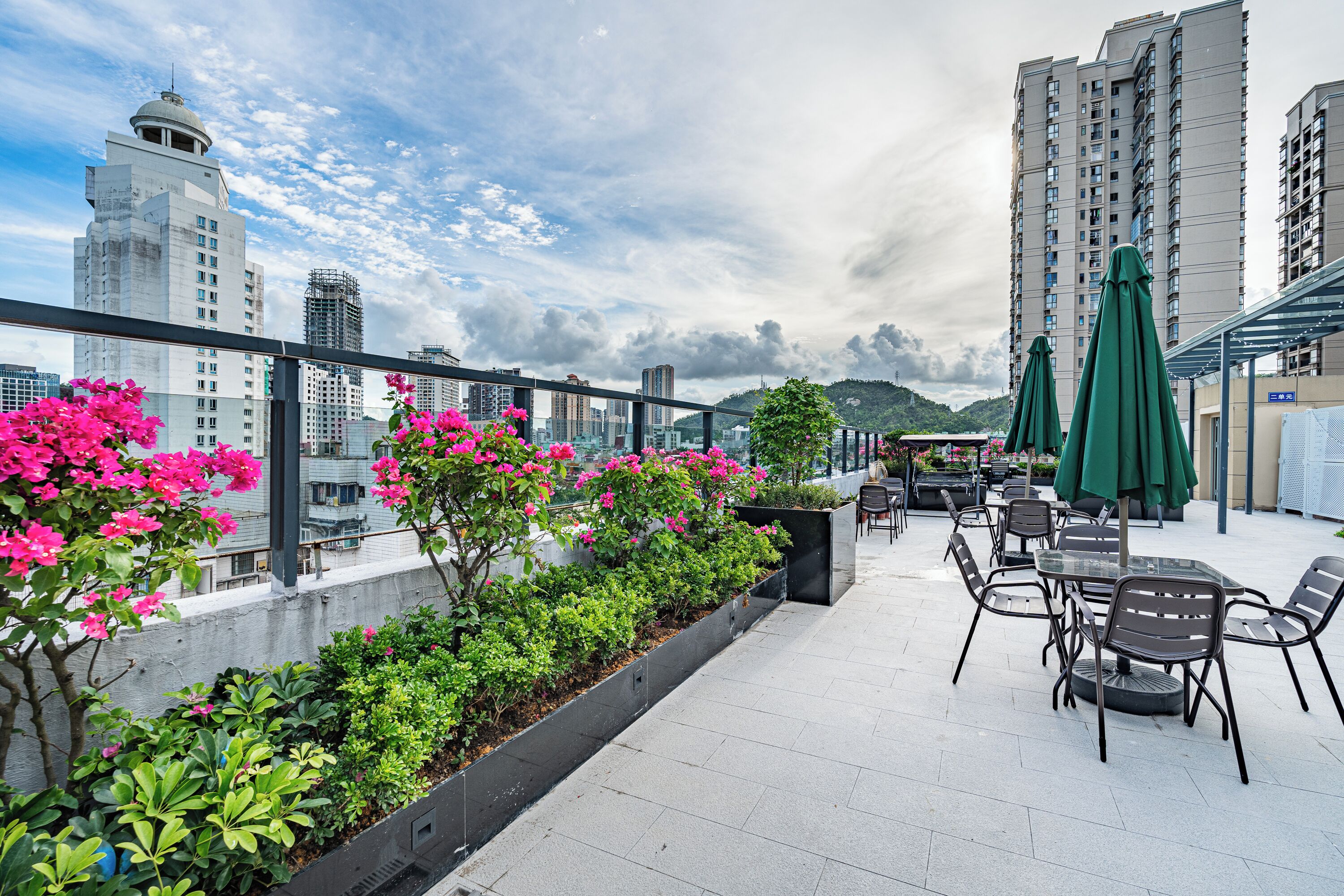 A rooftop terrace with a dining area, green umbrellas, and a view of city buildings.