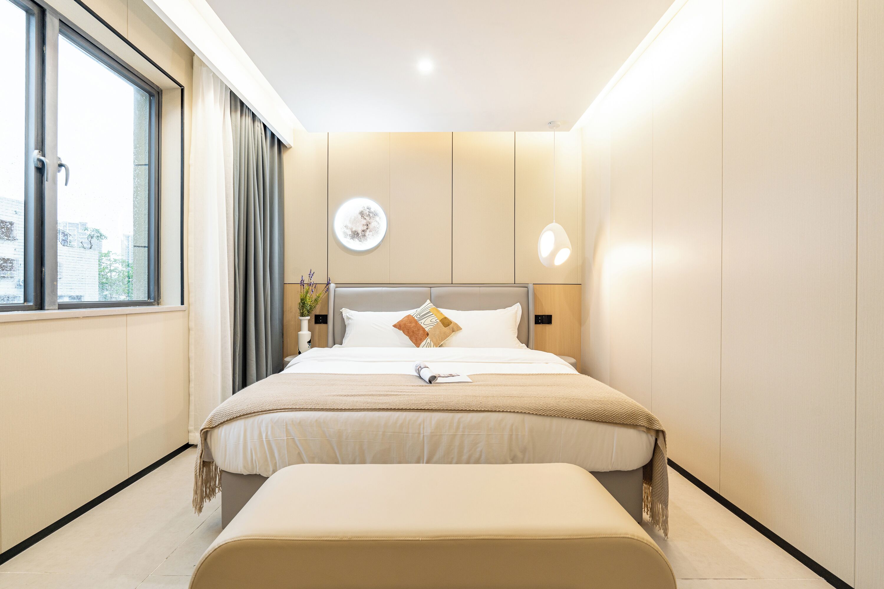 A modern bedroom with a large bed, a bedside table with a lamp, a window with curtains, and a wall-mounted light fixture.